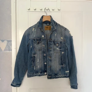 Blå jeansjacka med slitningar - Säljer en klassisk blå jeansjacka med slitna detaljer och knappar framtill. Jackan har två bröstfickor med lock och två sidofickor. Perfekt för dig som gillar en lite tuffare stil. Har aldrig använts. 