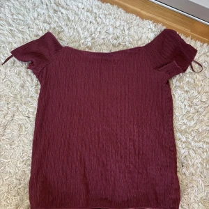 Vinröd offshoulder topp från 157 - Säljer en vinröd offshoulder topp från 157 i ett lätt skrynklat tyg. Toppen har korta ärmar med knytband och en luftig passform. Perfekt för varma dagar eller att styla med jeans.