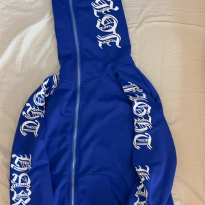 Blå hoodie med vit text - Säljer en blå hoodie med dragkedja och vit text i gotisk stil på ärmar och huva. Jackan har lång ärm och huva, perfekt för en avslappnad streetwear-look. Snygg och bekväm att bära till vardags. Dragkedjan går ända upp.