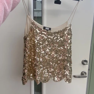 Guldig paljettopp med tunna axelband från Bik Bok - Säljer en glittrig guldig topp från Bik Bok med tunna axelband och paljetter över hela framsidan. Toppen har en vågig nederkant och är perfekt för att sticka ut. Baksidan är i ett slätt, beige tyg.
