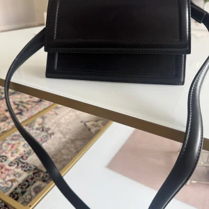 Svart axelväska i skinnimitation - Säljer en stilren svart axelväska med enkel design och magnetlås. Väskan har en lång axelrem och guldiga metalldetaljer. Perfekt för att bära det viktigaste när du är på språng. Helt oanvänd saknas bara prislapp.