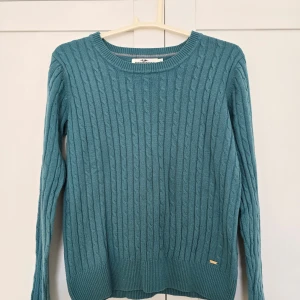 En turkos och en röd kabelstickad tröja  - Säljer en turkos och en röd kabelstickad tröja. Tröjan har långa ärmar och ribbade muddar vid ärmslut och nederkant. Klassisk och stilren modell med diskret logga nertill. Turkos tröjan är S och den röda XS