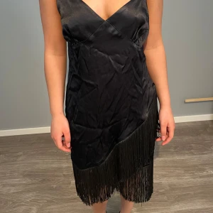 Svart klänning med fransdetaljer - Säljer en svart klänning i satinliknande material med smala axelband och v-ringad hals. Klänningen har fransar längs nederkanten och snedskuren fransdetalj framtill. Perfekt för dig som vill sticka ut med en elegant och trendig look.