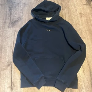 Axel Arigato Hoodie - Säljer en stilren mörkblå hoodie från Axel Arigato med liten vit logga på bröstet. Tröjan har huva och en stor magficka framtill. Perfekt för en avslappnad och trendig look.