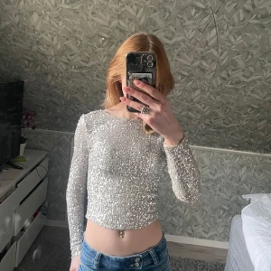 Glittrig silver topp med öppen rygg - Säljer en glittrig långärmad topp i silver med tight passform och öppen rygg. Toppen är croppad och har ett skimrande material som verkligen sticker ut. Perfekt för dig som vill glänsa lite extra!