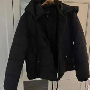 Svart pufferjacka med huva - Säljer en svart pufferjacka med huva och dragkedja framtill. Jackan har långa ärmar, två fickor med knappstängning och en lite boxig passform. Perfekt för kyligare dagar när du vill hålla dig varm och bekväm. Den är inte använd mer än 5 gånger, jag är 178 cm lång och den sitter bra på mig. Ordinarie pris 749kr.