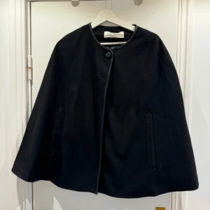 Svart cape från Zara Basic - Säljer en svart cape från Zara Basic med rund halsringning och knappstängning framtill. Capen har en stilren design med diskreta fickor och kantband i skinnimitation. Perfekt för dig som vill ha en elegant och tidlös look. Strl S!🥰