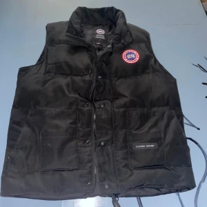 Svart dunväst från Canada Goose - Säljer en svart dunväst från Canada Goose med klassisk logotyp på bröstet och två stora fickor framtill. Västen har dragkedja och knappar samt hög krage. Perfekt för lager-på-lager under kyliga dagar. 