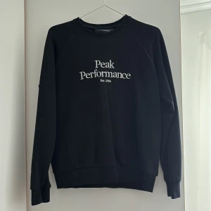 Svart sweatshirt från Peak Performance - Svart sweatshirt från Peak Performance med vit text på bröstet. Klassisk rund halsringning och ribbade muddar vid ärmslut och nederkant. Ett litet hål vid armen som är igen sytt, inget som syns. 