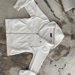 Vit hoodie från Polo Ralph Lauren - Vit hoodie från Polo Ralph Lauren med dragkedja och huva. Klassisk blå broderad logga på bröstet och två fickor framtill. Mjuk insida och ribbade muddar. Perfekt för en clean och sportig look den är liten i storlek.