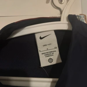 PSG Nike vindjacka navy S - Snygg navyblå PSG vindjacka från Nike med gul logga och Qatar Airways-tryck på bröstet. Jackan har dragkedja framtill, lång ärm och är tillverkad i lätt polyester med Dri-FIT-teknologi. Perfekt för dig som älskar fotboll och vill ha en sportig look.