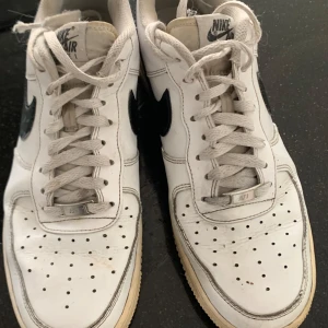 Nike Air Force 1 vita sneakers - Klassiska Nike Air Force 1 sneakers i vitt skinn med svart swoosh. Skorna har snörning, perforerad tå och platt sula.  Mer kvar att ge