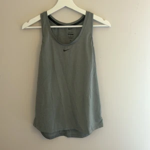 Grå Nike Dri-Fit linnetopp M - Säljer en grå linnetopp från Nike i modellen Dri-Fit. Toppen har en sportig look med brottarrygg och svart Nike-logga framtill. Materialet är lätt och andas, perfekt för träning eller gym. Slim fit-passform som sitter snyggt på kroppen.
