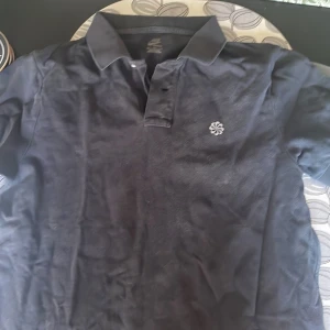 Svart pikétröja från Nike - Svart pikétröja från Nike med klassisk krage och korta ärmar. Tröjan har en vit broderad blomliknande logga på bröstet och är tillverkad i bomull. Perfekt för en sportig och stilren look.