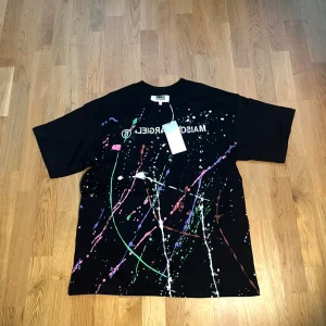 Maison Margiela svart t-shirt S - Svart t-shirt från Maison Margiela MM6 med färgstänk i grönt, rosa, vitt, lila och orange. Oversized fit, korta ärmar och tryckt logga på bröstet. Tillverkad i mjuk bomull, perfekt för dig som gillar statement-plagg.