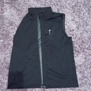 Svart väst från Pelle P i storlek L helt ny inte ens provat - Svart väst från Pelle P med hög krage och hel dragkedja framtill. Västen har diskret logga på bröstet och ryggen, samt meshfoder på insidan. Perfekt för lager-på-lager och sportiga outfits.