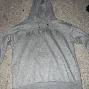 Grå hoodie med strass Ma Chérie - Grå hoodie med huva och snörning, dekorerad med texten 'MA CHÉRIE' i strass på bröstet. Storleken är 134/140. Mjuk och skön med normal passform och långa ärmar. Perfekt för dig som gillar en chill och snygg stil.