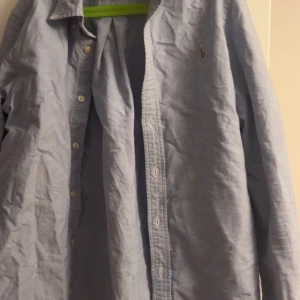 Blå skjorta från Ralph Lauren - Klassisk ljusblå skjorta från Ralph Lauren med broderad logga på bröstet. Skjortan har lång ärm, knappar framtill och är tillverkad i bomull. Perfekt för en clean och stilren look.