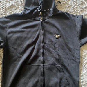 Svart hoodie med dragkedja från Prada - Svart hoodie från Prada med hel dragkedja och klassisk huva. Tröjan har en snygg Prada-logga i metall på bröstet och stora fickor framtill. Tillverkad i mjuk bomull, perfekt för en clean och stilren look.