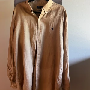 Beige linneskjorta från Polo Ralph Lauren - Snygg beige skjorta från Polo Ralph Lauren i linne med klassisk button-down krage och broderad logga på bröstet. Skjortan har lång ärm och custom fit-passform, perfekt för en stilren och avslappnad look.
