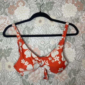 Orange blommig bikini med knyt - Snygg bikini i orange med stora vita blommor och tropiskt mönster. Överdelen har breda axelband och en söt knytning baktill. Underdelen är högt skuren och matchar med samma blommiga print. Perfekt för strandhäng och sommarvibbar!