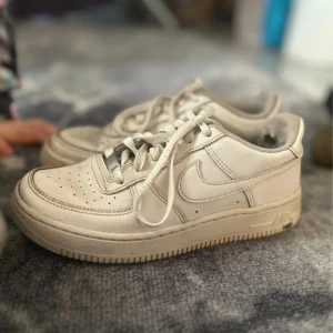 Nike Air Force 1 vita sneakers - Klassiska Nike Air Force 1 sneakers i helvitt skinn med rund tå och platt sula. Skorna har perforerade detaljer på tån, snörning och den ikoniska swoosh-loggan på sidorna. Perfekta för en clean och tidlös streetstil.