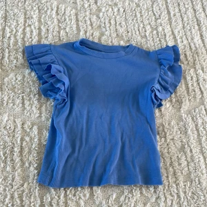 Blå volangtopp från Gina Tricot - Supersöt blå topp från Gina Tricot med ribbad struktur och korta ärmar med stora volanger. Perfekt för dig som vill ha något extra till outfiten. Rund halsringning och mjukt material som känns skönt mot huden.