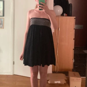 Svart axelbandslös kortklänning - Svart axelbandslös kortklänning med smockad överdel i vitt broderat mönster. Klänningen har en luftig och flowy passform, perfekt för varma dagar. Den är i storlek M och kan användas både som klänning samt kjol. 