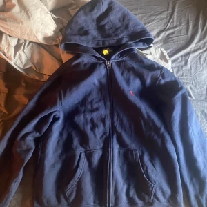 Polo hoodie - Marinblå hoodie från Polo Ralph Lauren med dragkedja och huva. Klassisk röd broderad logga på bröstet och två fickor framtill. Tillverkad i mjuk bomull, perfekt för chill dagar eller när du vill ha en clean look.. de är en storlek xl i barn size sitter som en small