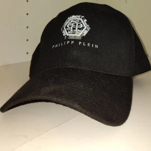 Svart keps från Philipp Plein - Stilren svart keps från Philipp Plein med böjd skärm och vit logotyp framtill. Klassisk passform och tillverkad i ett slitstarkt material som ger en cool och sportig look. Perfekt accessoar för att lyfta din outfit.