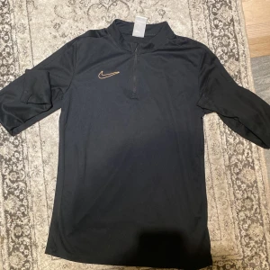 Svart Nike Dri-Fit träningströja XL - Svart Nike Dri-Fit träningströja i funktionsmaterial med halv dragkedja och guldigt broderat swoosh på bröstet. Tröjan har lång ärm och sportig passform, perfekt för dig som gillar att träna och vill ha en stilren look.