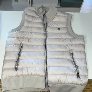 Beige dunväst från Burberry - Snygg beige dunväst från Burberry med klassisk TB-logga på bröstet. Västen har quiltad design, ribbade muddar och två fickor med dragkedja. Perfekt för lager-på-lager och stilren look. Innerfoder i satin och diskret Burberry-märke på insidan jätte skönt och fint. Finns i fler storlekar 