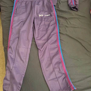 Palm angles byxor - Säljer en lila tracksuit från Palm Angels i storlek M. Setet har vita dragkedjor, logga på både jacka och byxa samt ränder i rosa och blått längs ärmar och ben. Materialet är mjukt och syntetiskt, perfekt för en sportig look. 
