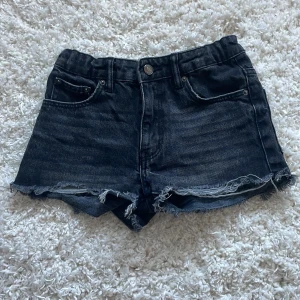 Svarta jeansshorts från Gina Tricot - Snygga svarta jeansshorts från Gina Tricot x Iza & Elle med råa, fransiga kanter och klassisk femficksmodell. Shortsen har hög midja och en cool, lite tvättad look som passar perfekt till sommaren. Styla med en crop top eller hoodie för en chill vibe.