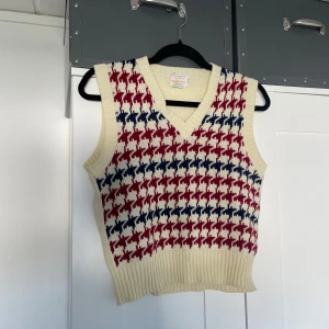Stickad väst med röd och blå hundtandsmönster - Retro stickad väst från domkenny i krämvitt med hundtandsmönster i rött och mörkblått. V-ringad och ribbstickad vid ärm och nederkant, passar både M och L!