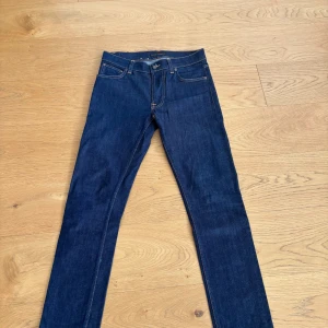 Mörkblå skinny jeans från Nudie Jeans - Snygga mörkblå skinny jeans från Nudie Jeans med klassiska kontrastsömmar och femficksdesign. Tillverkade i 100% ekologisk bomull. Bakfickorna har Nudie Jeans signaturbrodyr och läderpatch i midjan. Perfekta för dig som gillar stilrena och tidlösa jeans.