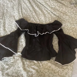 Svart offshoulder blus från Zara - Svart offshoulder blus från Zara Basic med bred volang runt axlarna och vida trumpetärmar. Toppen har vita kanter längs volangerna och ärmarna för en snygg kontrast. Perfekt för dig som gillar statement-ärmar och en trendig look.