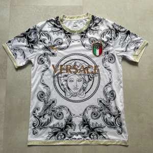 Versace tröja - Snygg vit fotbollströja med svart barockmönster, guldiga detaljer och Versace-tryck på bröstet. Italiens landslagsemblem på vänster sida och Puma-logga i guld på höger. Guldmönstrad kant vid ärmar och hals. Perfekt för dig som gillar unik stil.