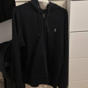 Svart hoodie från Ralph Lauren - Svart hoodie från Ralph Lauren med dragkedja framtill och klassisk huva med snörning. Liten broderad logga på bröstet. Perfekt för en chill och stilren look. Långärmad och i mjukt material som känns skönt mot huden.