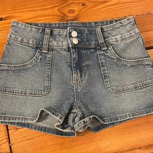 Högmidjade jeansshorts från H&M Divided - Snygga blå jeansshorts från H&M Divided med hög midja och dubbla knappar framtill. Klassisk femficksmodell med breda bälteshällor och ljus tvätt. Perfekta för varma dagar och enkel att matcha med allt.