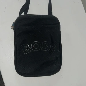 Svart axelväska från BOSS - Snygg svart axelväska från BOSS med stilren design och logga framtill. Väskan har justerbar rem, dragkedja med metall-detalj och är tillverkad i slitstark polyester. Perfekt storlek för mobil, plånbok och andra småsaker.