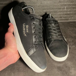 Svarta Saint Laurent sneakers - Svarta sneakers från Saint Laurent med vit logga broderad på sidan. Skorna är i storlek 43. Skick är mycket bra 9/10. OG/tillbehör inget därav det lägre priset. Givetvis är dom ÄKTA vilket garanterar. Obs sulkräm har använts 