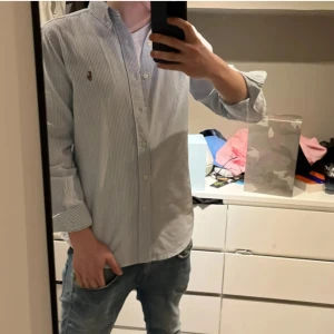 Randig skjorta från Polo Ralph Lauren - Klassisk skjorta från Polo Ralph Lauren i vitt och ljusblått med smala ränder. Skjortan har lång ärm, knappar hela vägen och broderad logga på bröstet. Tillverkad i bomull och har en normal passform med button-down krage. 