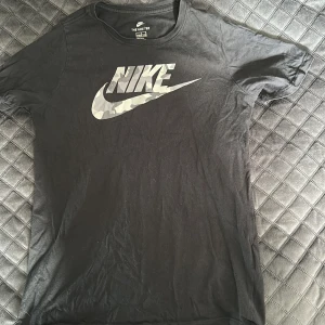 Svart Nike t-shirt med camologga - Svart t-shirt från Nike med klassisk rund hals och korta ärmar. Stor Nike-logga i grått och vitt camomönster på bröstet. Tillverkad i mjuk bomull, perfekt för en sportig och avslappnad stil.