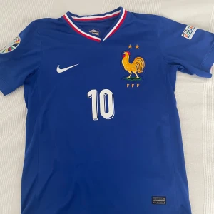 Frankrike Mbappé 10 Nike fotbollströja - Blå officiell Frankrike fotbollströja från Nike med Mbappé och nummer 10 på ryggen. Tröjan har korta ärmar, broderad tupp-logga, två stjärnor och Dri-FIT-material. Kragen har röd, vit och blå kant. Perfekt för dig som älskar landslagsfotboll.