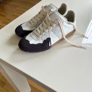 Maison Margiela gats paint splatter - Maison Margiela sneakers med unik design i vitt och svart. Skorna har beige snörning, detaljer i mocka och läder samt en cool kontrast mellan ljusa och mörka partier. Sulan är i gummi med klassiskt Margiela-tryck undertill. Perfekt för dig som gillar statement-sneakers.