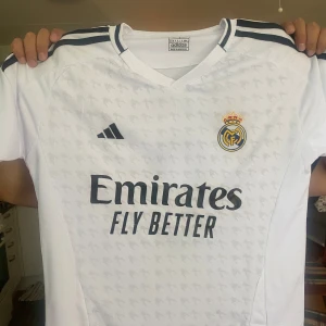 Real Madrid Mbappé 9 Adidas tröja - Snygg vit Real Madrid fotbollströja från Adidas med Mbappé och nummer 9 på ryggen. Tröjan har svarta detaljer, klubbmärke på bröstet och Emirates Fly Better-tryck. Tillverkad i lätt och ventilerande material, perfekt.