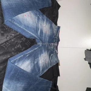 Dsquared2 blå jeans med slitningar - Säljer ett par blå Dsquared2 jeans med coola slitningar, färgstänk och distressed detaljer. Jeansen har en klassisk femficksmodell, normal passform och är tillverkade i jeansmaterial. Perfekt för dig som gillar en edgy streetstil.