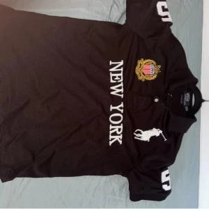  Polo Ralph Lauren chief keef - Svart pikétröja från Polo Ralph Lauren med stora vita broderier, New York-tryck och ikonisk polospelare på bröstet. Snygga siffror 5 på ärmar och rygg, samt guldfärgat broderat emblem. Klassisk krage och korta ärmar. Perfekt för en sportig och stilren look.