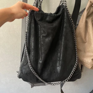 Stella mccartney liknande väska - Snygg svart axelväska i mocka-look med glansig finish och kraftiga silverfärgade kedjor som axelremmar. Väskan har en mjuk, slouchy form och stängs med snörning upptill. Perfekt för dig som gillar statement-accessoarer. Bra skicka förutom att två sömmar släppt men de är inget som syns när man bär den❤️❤️